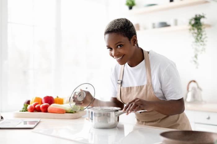 Smiling young african american woman in apron prepare eat and smell dish in minimalist kitchen interior