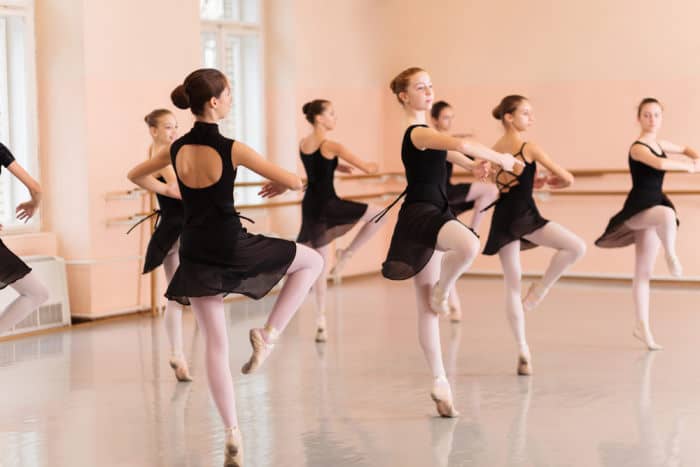 Medium group of teenage girls in black dresses practicing ballet moves in a large dance studio. Exercising pivot turns, standing on one leg
