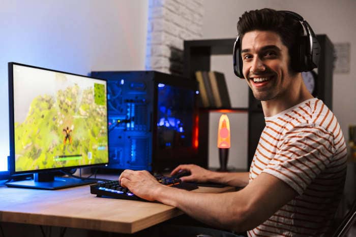 Portrait of happy smiling guy playing video games on computer wearing headphones and using backlit colorful keyboard